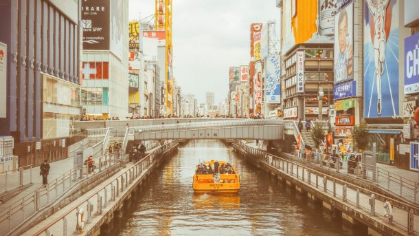 An aged photo of a water taxi going under a bridge with Osaka, Japan city surrounding